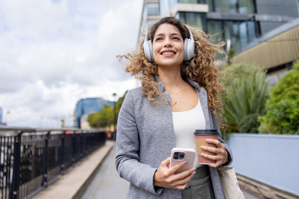 woman walking holding her coffee and listening to a podcast with headphones on