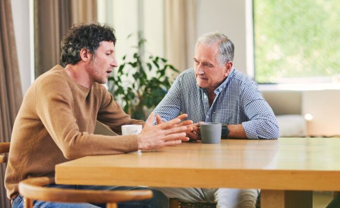 Two men sitting at a table having a serious conversation over coffee, with one man speaking and gesturing while the other listens attentively.