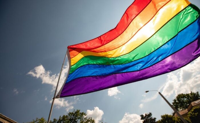 A rainbow Pride flag waves in the wind against a sunny blue sky with scattered clouds, trees, and a streetlight visible in the background.