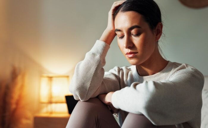 A young woman sits indoors with her eyes closed, resting her head on her hand. She looks distressed or exhausted. A warm lamp glows in the background.
