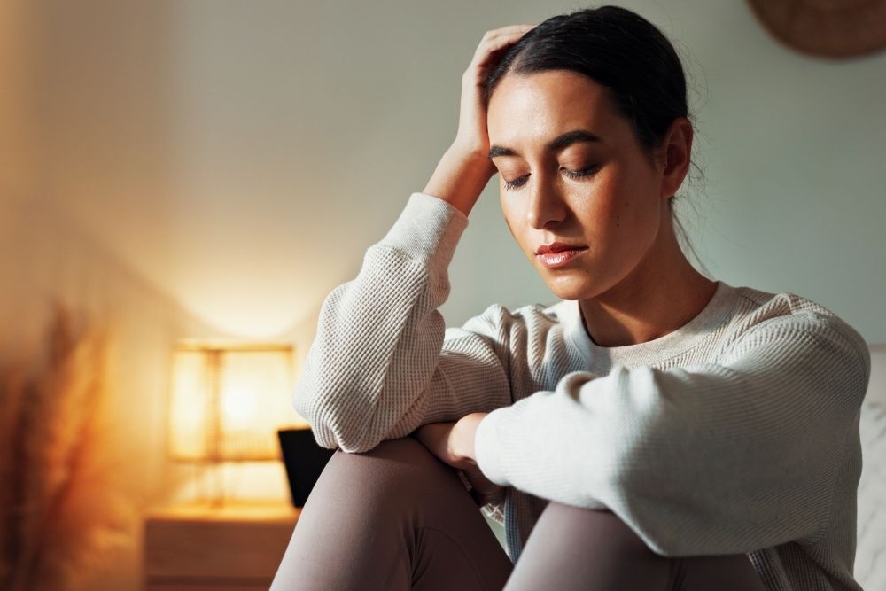 A young woman sits indoors with her eyes closed, resting her head on her hand. She looks distressed or exhausted. A warm lamp glows in the background.