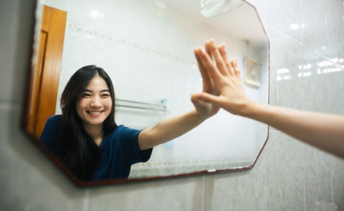 A smiling woman in a bathroom gently touches her reflection in the mirror, symbolizing self-acceptance, confidence, and positive self-talk.