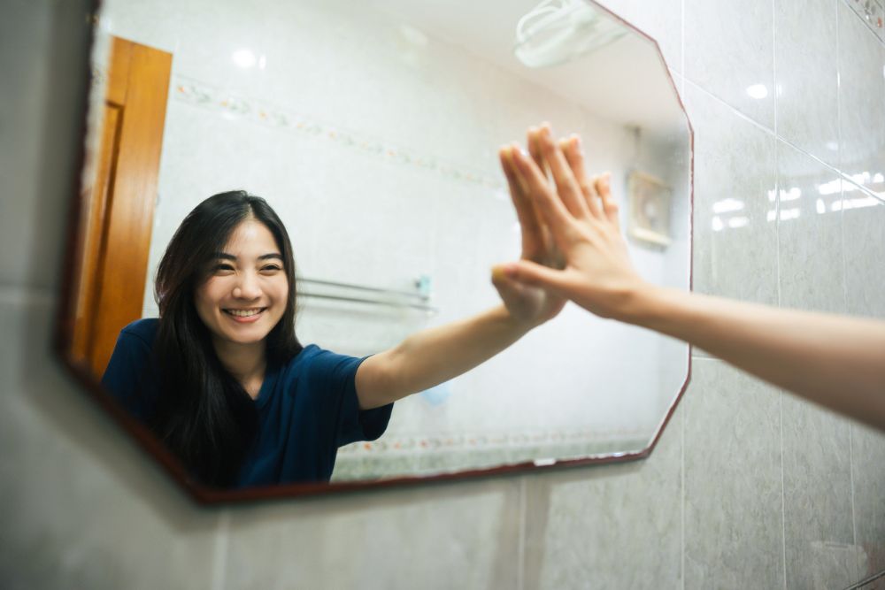 A smiling woman in a bathroom gently touches her reflection in the mirror, symbolizing self-acceptance, confidence, and positive self-talk.