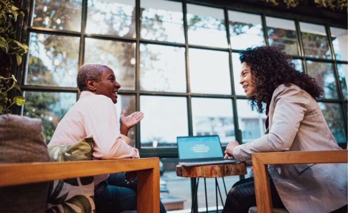 Two women sitting across from each other in a cozy café, engaged in a supportive conversation, with a laptop open on the table between them.