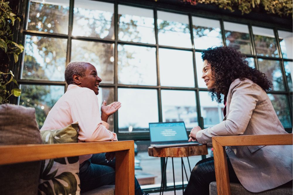 Two women sitting across from each other in a cozy café, engaged in a supportive conversation, with a laptop open on the table between them.