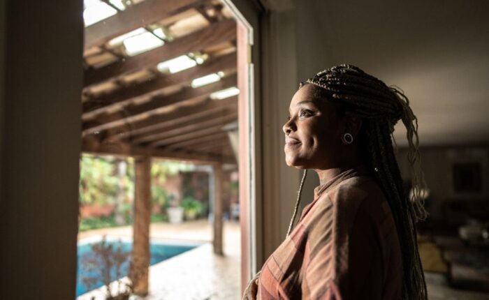 Woman with braided hair standing indoors, looking out a window with a calm and hopeful expression, symbolizing reflection, healing, and recovery.