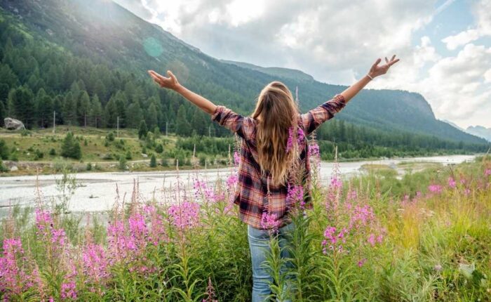 A woman stands in a field of wildflowers with arms raised, facing mountains and a river, symbolizing freedom, purpose, and connection with nature.