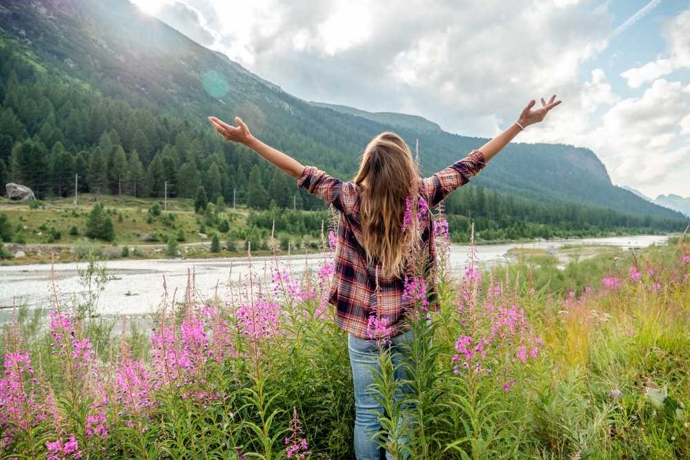 A woman stands in a field of wildflowers with arms raised, facing mountains and a river, symbolizing freedom, purpose, and connection with nature.