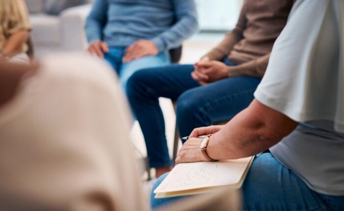 Close-up of people sitting in a therapy group session, with one person holding a notebook and others sitting in a circle, suggesting discussion and support.