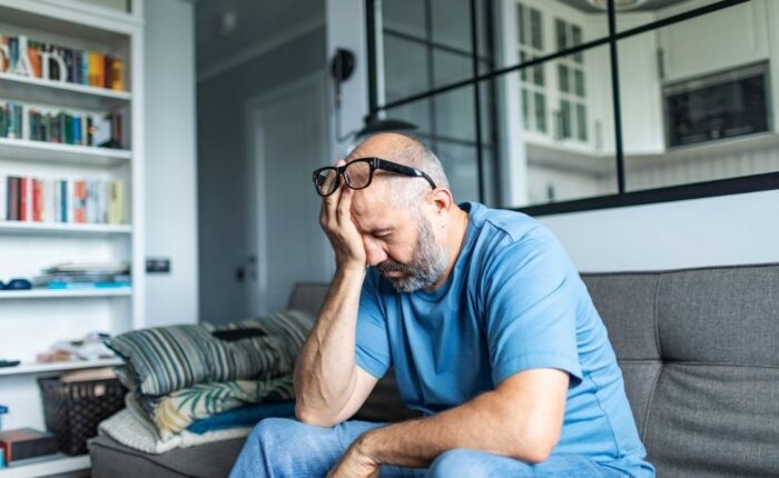 Middle-aged man sitting on a couch with his hand on his face, looking stressed and overwhelmed in a home setting