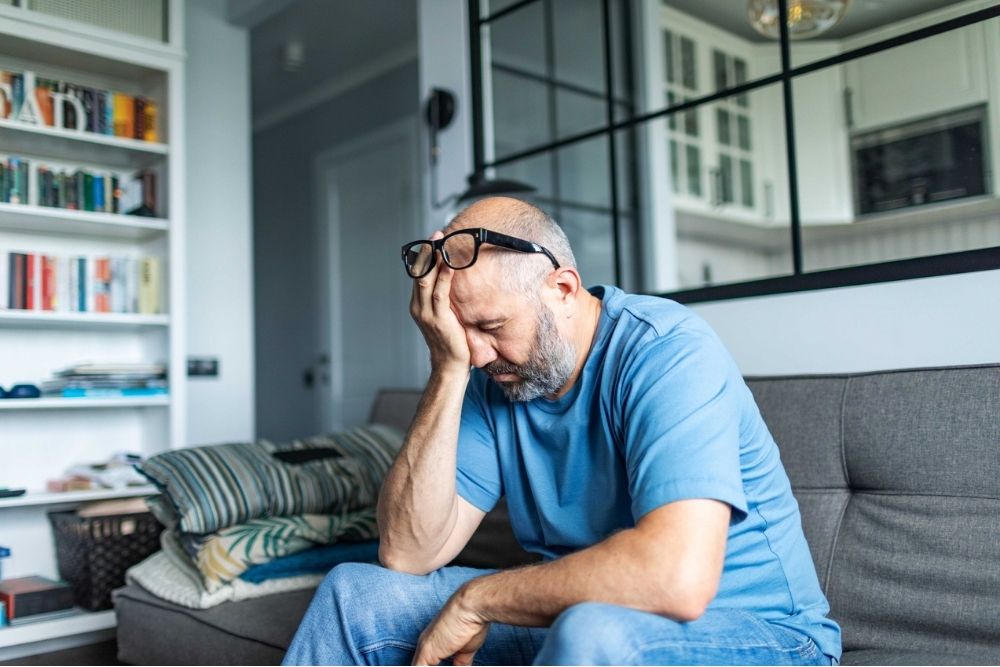 Middle-aged man sitting on a couch with his hand on his face, looking stressed and overwhelmed in a home setting