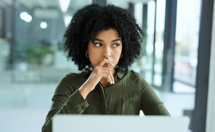 A woman sits indoors looking thoughtful and concerned, with her hand resting near her mouth, suggesting anxiety or deep thinking.