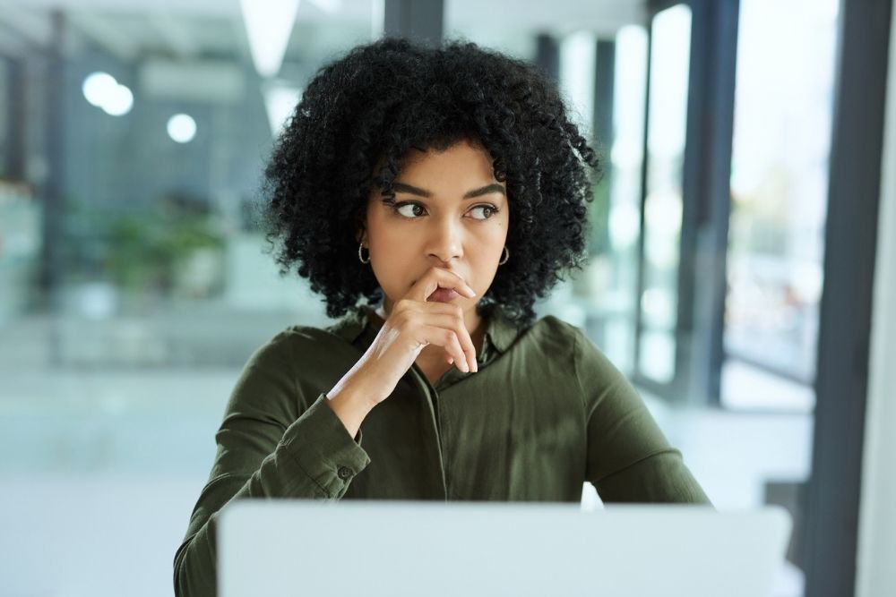A woman sits indoors looking thoughtful and concerned, with her hand resting near her mouth, suggesting anxiety or deep thinking.