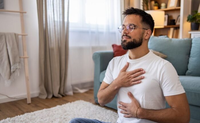 Man sitting on the floor in a relaxed position with eyes closed, practicing mindful breathing with hands on chest and abdomen in a cozy living room.