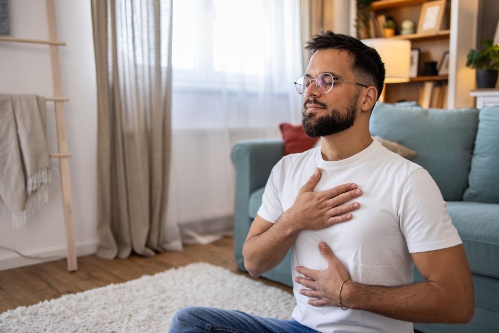 Man sitting on the floor in a relaxed position with eyes closed, practicing mindful breathing with hands on chest and abdomen in a cozy living room.