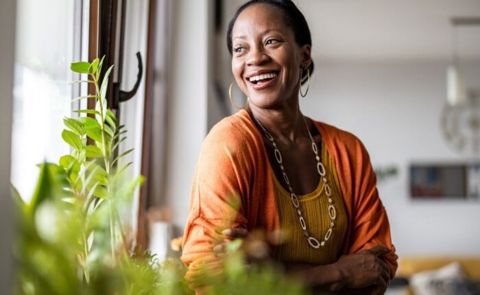 A woman stands by a window surrounded by green plants, smiling brightly. She wears an orange cardigan and a yellow top, appearing joyful and relaxed in a warm, inviting indoor space.