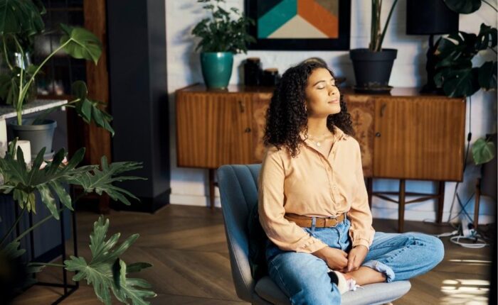 Young woman sitting cross-legged in a cozy living room, eyes closed and breathing calmly while practicing mindfulness surrounded by houseplants.
