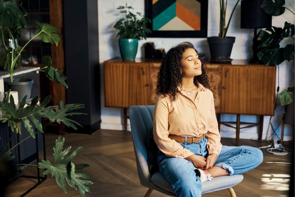Young woman sitting cross-legged in a cozy living room, eyes closed and breathing calmly while practicing mindfulness surrounded by houseplants.