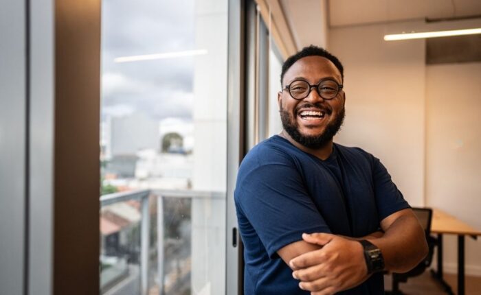 A smiling man standing indoors near large windows, arms crossed, appearing confident and happy.