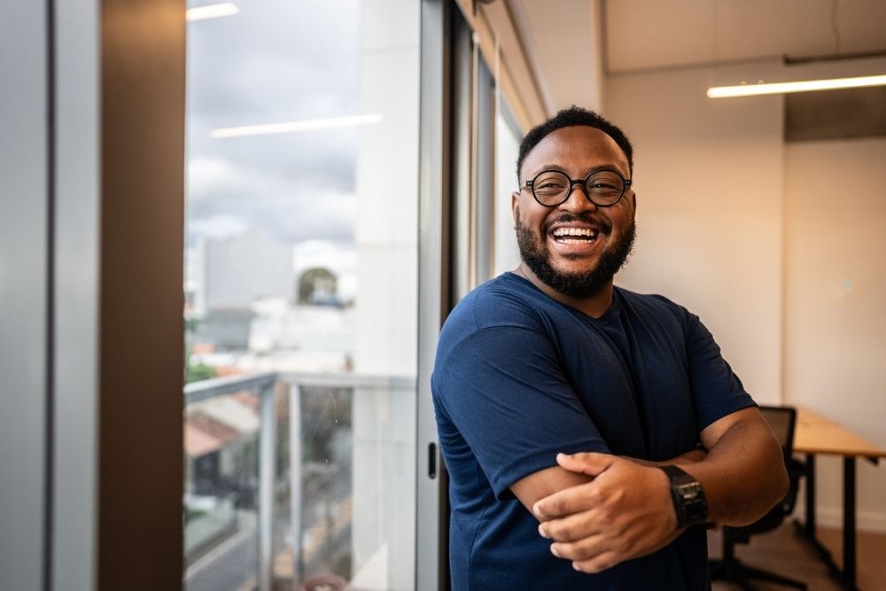 A smiling man standing indoors near large windows, arms crossed, appearing confident and happy.