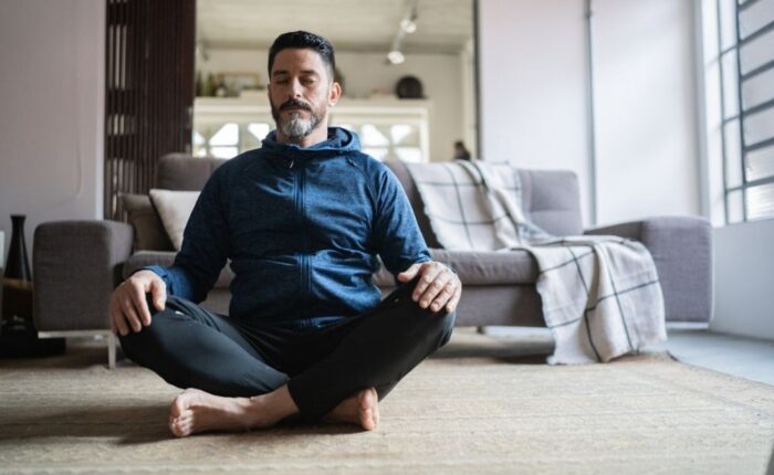 A man sits cross-legged on the floor with his eyes closed, practicing mindfulness or meditation in a calm living room.