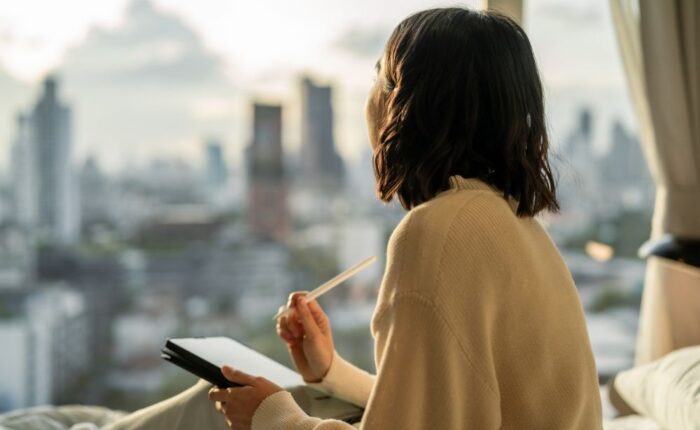 A person sits by a window overlooking a city skyline, holding a notebook and pen while reflecting quietly in a calm, sunlit room.