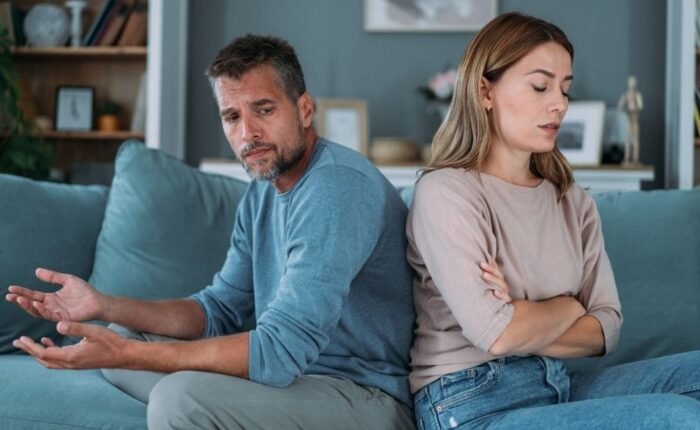 A man and woman sit back-to-back on a sofa, both with serious expressions. The woman has arms crossed, while the man gestures with open palms, conveying tension. Picture depicts a opportunity to find some positive ways to approach conflict