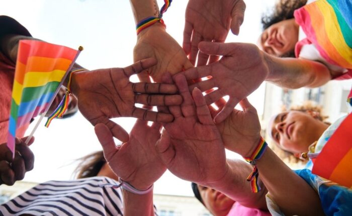 A group of multiracial people with rainbow LGBTQ bracelets stacking hands. Scene is one of unity and support for the LGBTQ community.